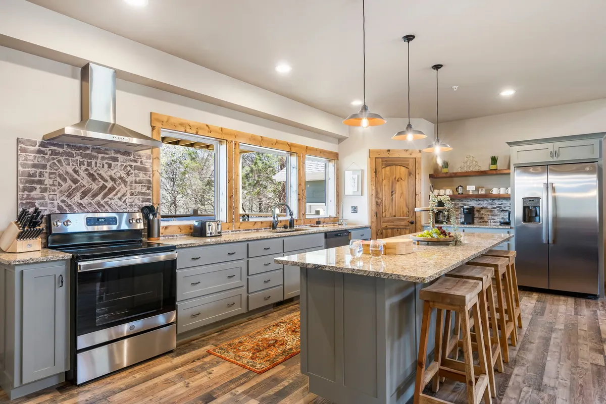 A close, bright view of the kitchen area reveals a central island with granite countertops, stainless steel appliances, large windows flooding the space with light, and warm wooden accents. The scene suggests a place for joyful meal prep and casual conversations.
