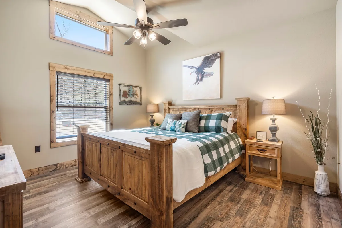 A cozy bedroom with a wooden bed frame, plaid bedding, and a window offering natural light. The room features an eagle artwork above the bed and blinds that can be adjusted for privacy or views. The atmosphere is serene, inviting relaxation and quiet moments.