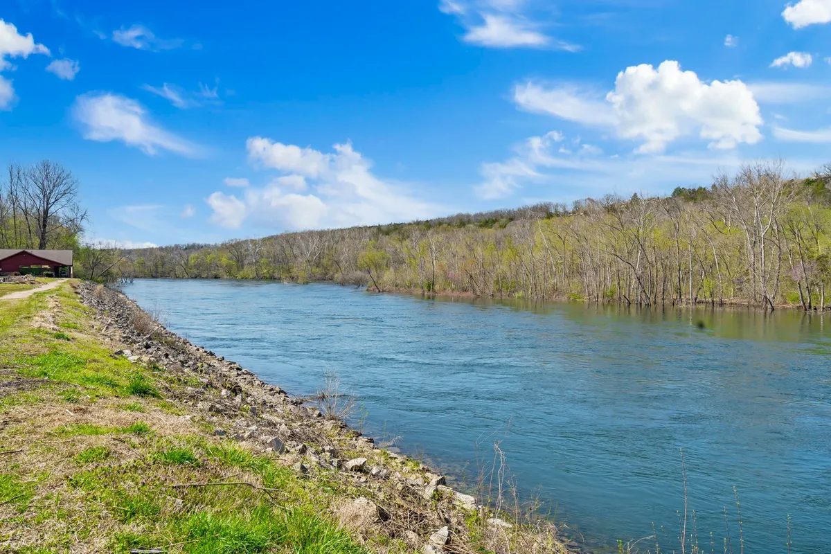 A picturesque view featuring a river bordered by dense trees and distant hills, with a pathway running alongside the water. The scene is serene, ideal for nature walks, birdwatching, and appreciating sunset reflections on the water.