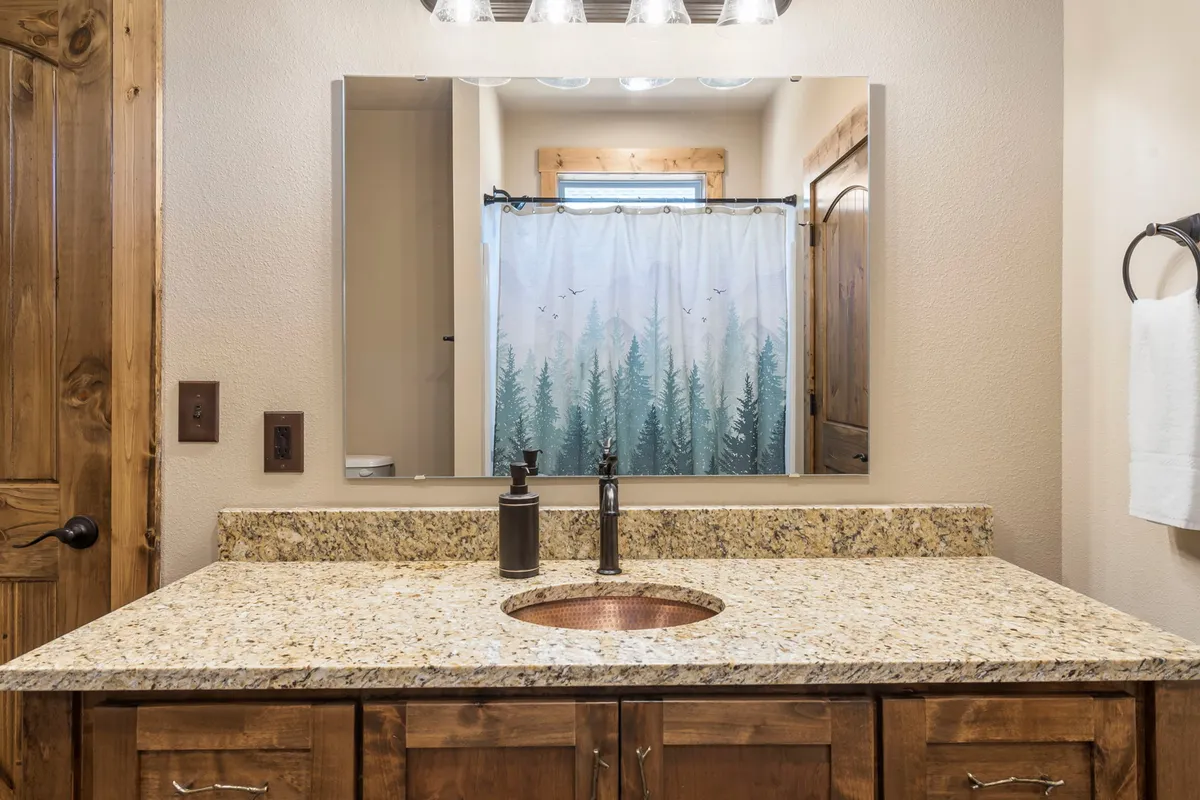 A serene bathroom with a granite countertop, copper sink, large mirror, and a forest-themed shower curtain. The space is calming, ideal for morning routines or relaxing after a busy day.