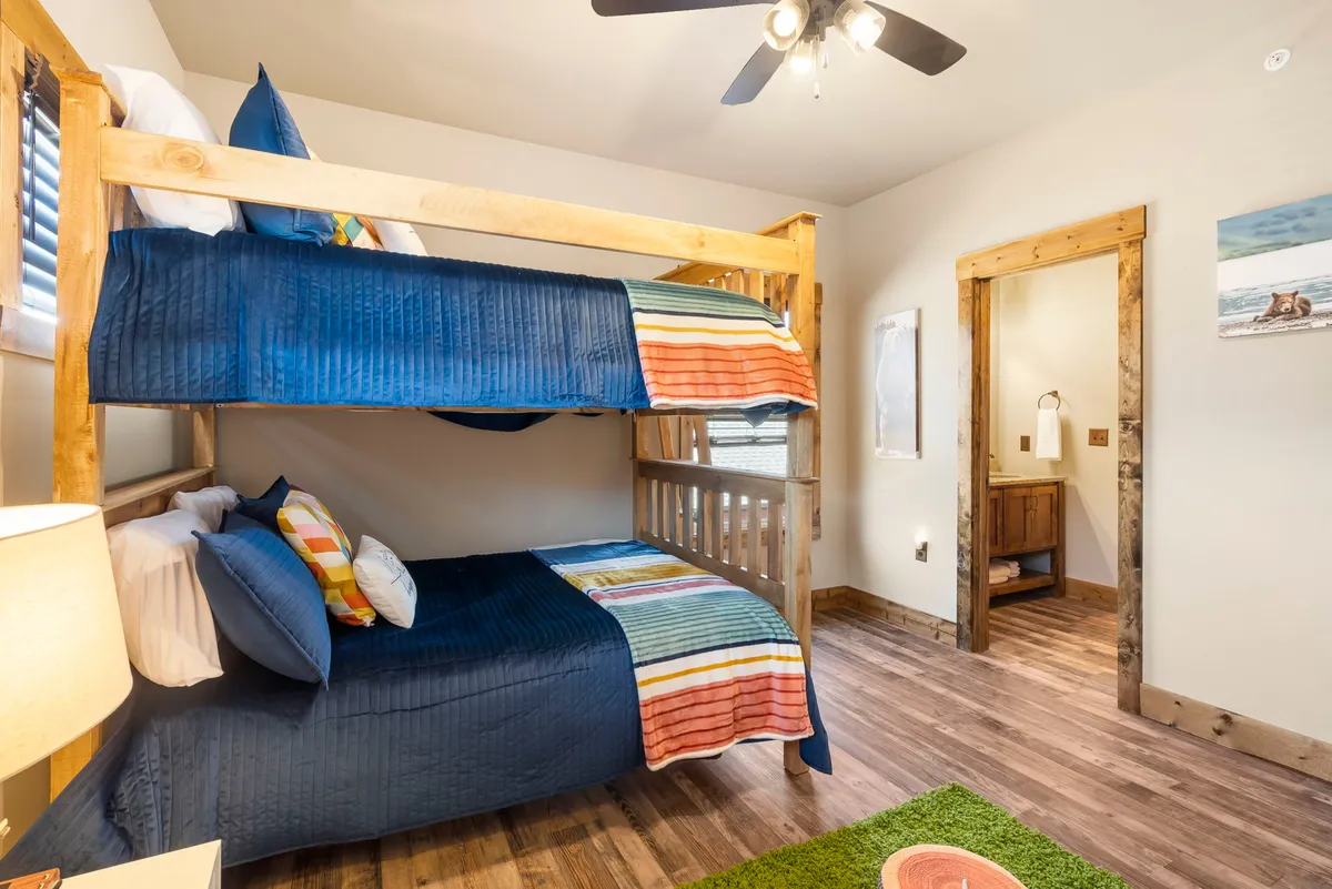 A cheerful bunk room with multiple beds dressed in colorful bedding, wooden floors, and a warm lamp. The space feels playful and relaxed—perfect for kids or groups wanting a fun sleepover vibe after a night of games.