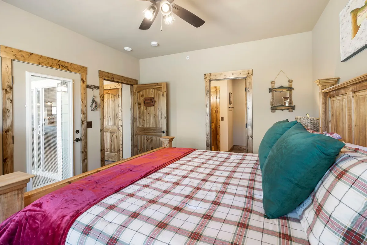 A comfortable bedroom with a plaid bedspread, colorful pillows, wooden furniture, and a doorway leading to an outdoor space. Morning sunlight filters through, creating a warm and inviting atmosphere.