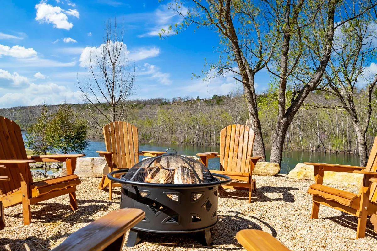 An outdoor fire pit area framed by Adirondack chairs, overlooking a tranquil water view and surrounded by trees. The scene invites campfire gatherings, storytelling, and starlit evenings, extending the warmth of the indoor space out to nature.