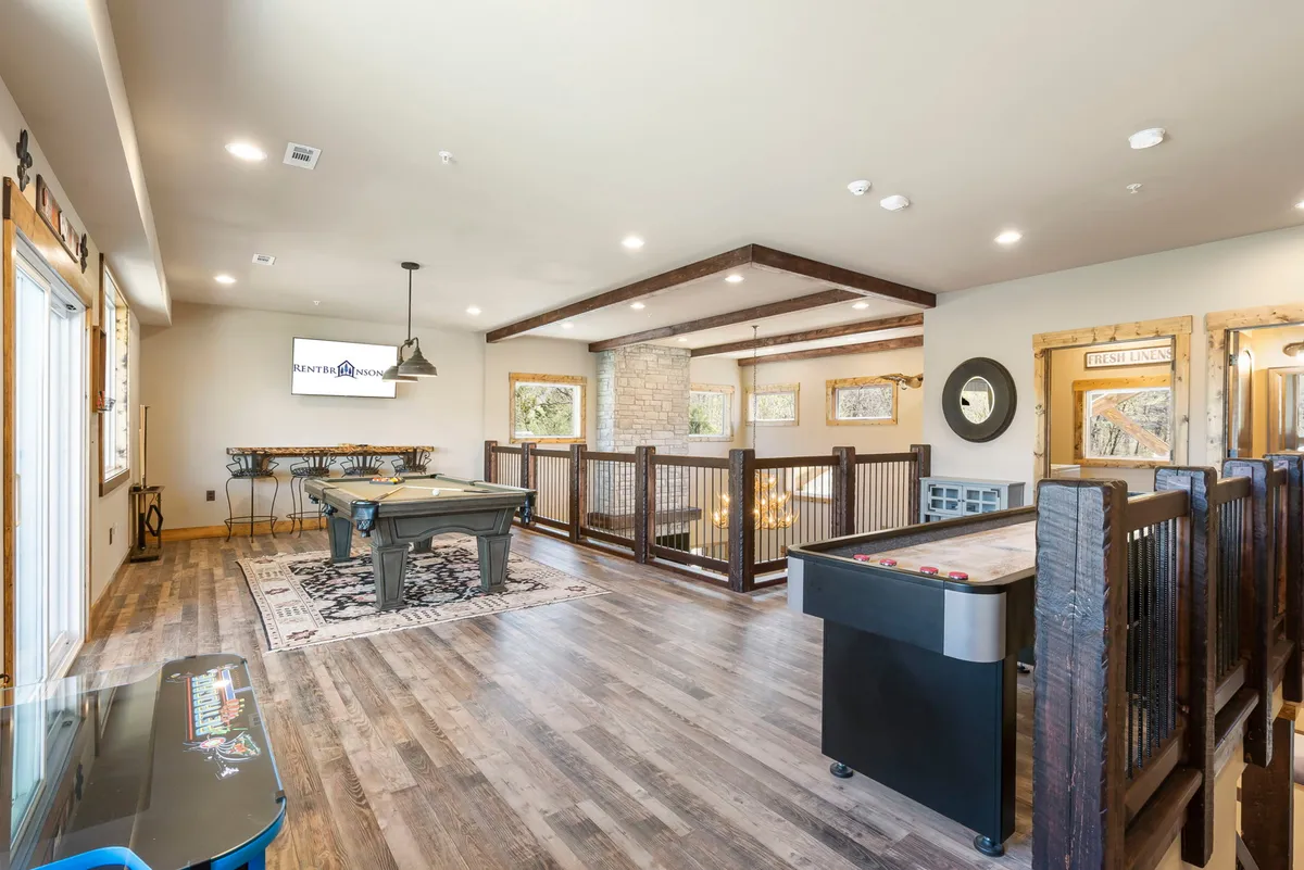 A spacious and well-lit game room featuring a pool table and air hockey table at its center. Large windows allow natural light to flood the space, and a chandelier adds an elegant touch. There's also a TV screen, providing multiple entertainment options. The overall vibe is energetic yet relaxed, making it perfect for group fun and gatherings.