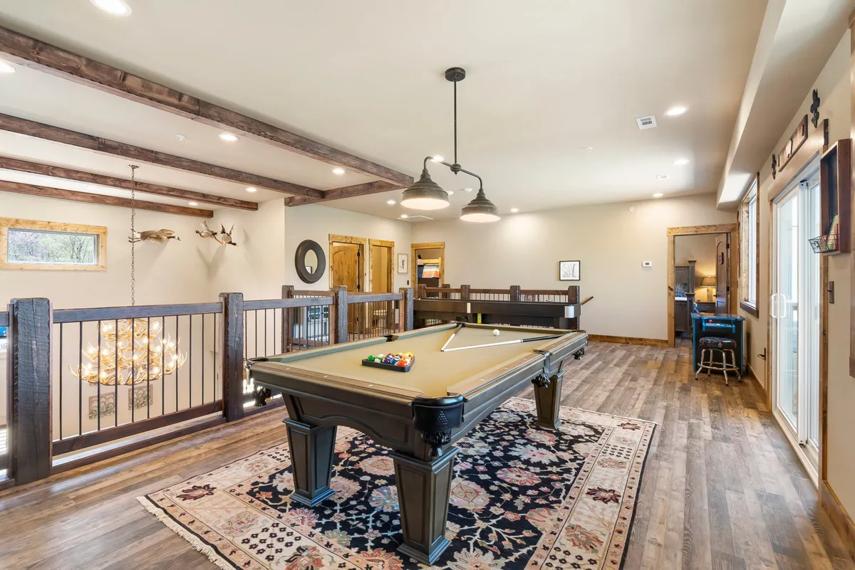 A vibrant interior game room featuring a stylish pool table beneath a chandelier, wooden beams overhead, and a cozy rug underfoot. The space is set up for socializing, friendly competition, and shared laughter during game nights.