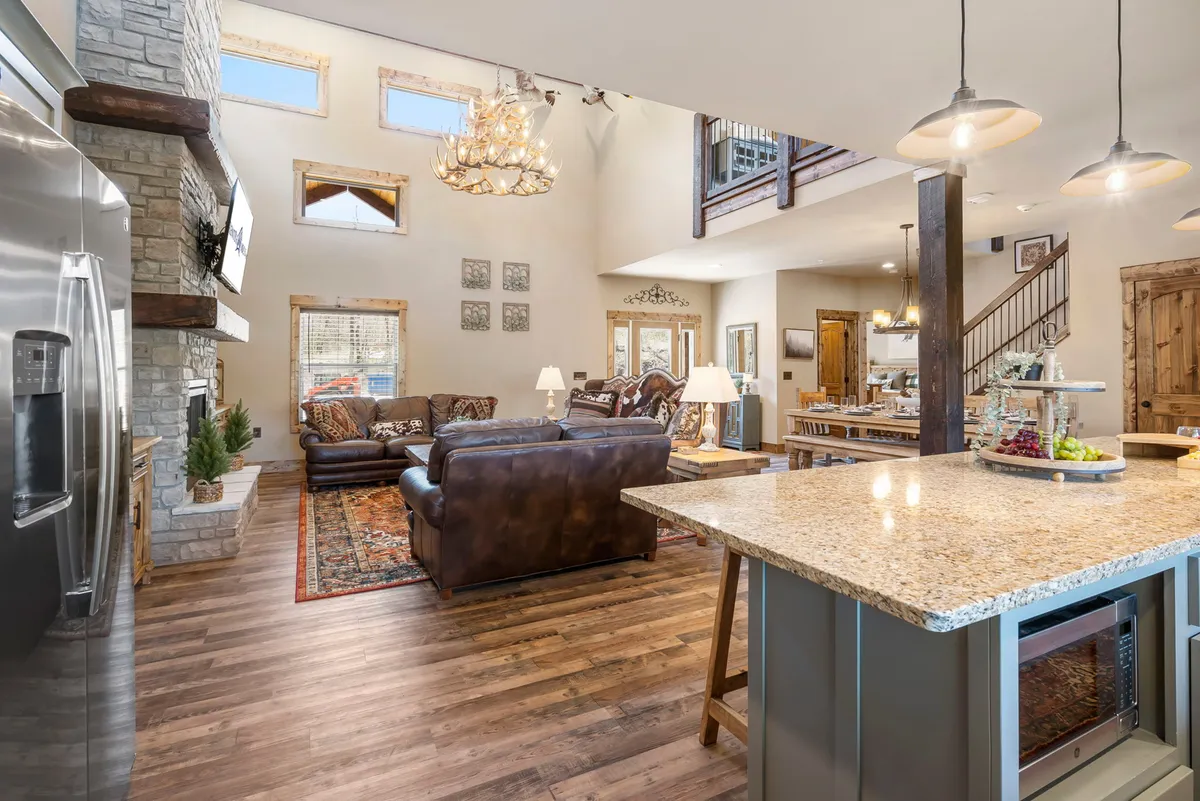 This image showcases a spacious open-concept area featuring a granite kitchen island, a striking stone fireplace, comfortable seating, and an elegant chandelier. The scene exudes warmth and invites gatherings, with visible zones for both relaxing and culinary activities.